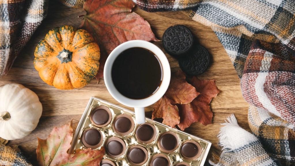 A cup of coffee on a wooden table surrounded by a fall blanket and autumn leaves.