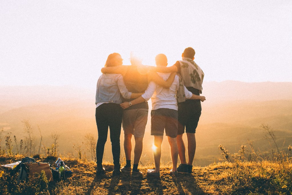 Four people  from behind with their arms around each other watching the sunrise.