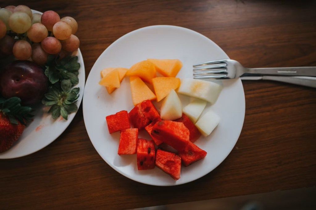 A white plate on a wooden table top with mixed fruit chunks and a fork on it.