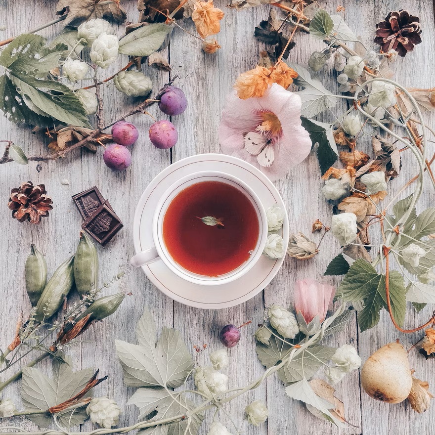 A cup of hot tea on a white table with flowers and herbs scattered around it.