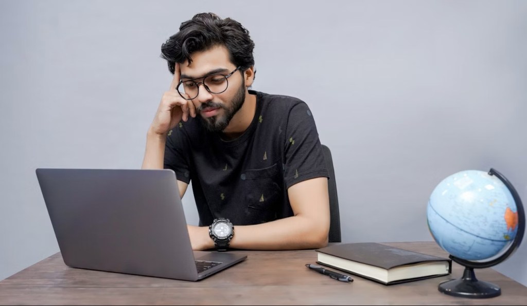 Man sitting at a table looking at a laptop with a book and small globe beside it.