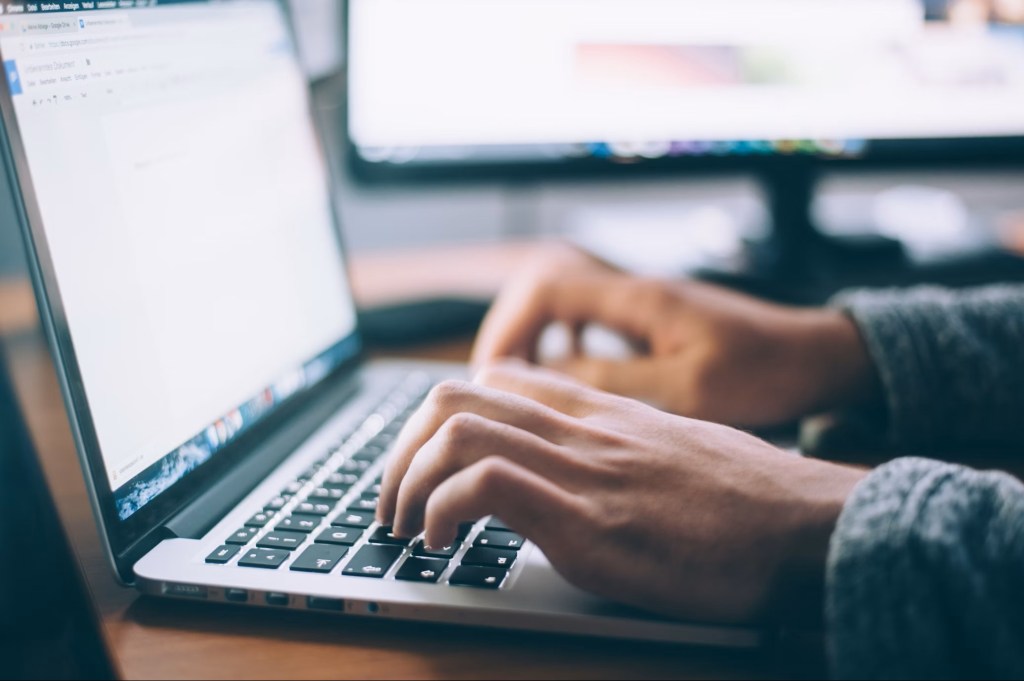Laptop on table with hands typing and monitor in background.