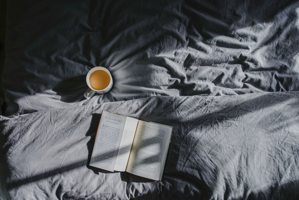 A rumpled bed in shadow with an open book and cup of tea on it.